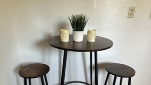 Ein kleiner runder Tisch mit drei Hocker, jeweils mit dunkel hölzernen Sitzflächen und schwarzen Metallbeinen. Auf dem Tisch steht eine weiße Vase mit grünen Pflanzen und zwei weiße Kerzenständer.