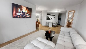 Moderni olohuone, jossa on suuri litteänäyttöinen televisio, kulmasohva, sohvapöytä ja taustalla ruokailutila.