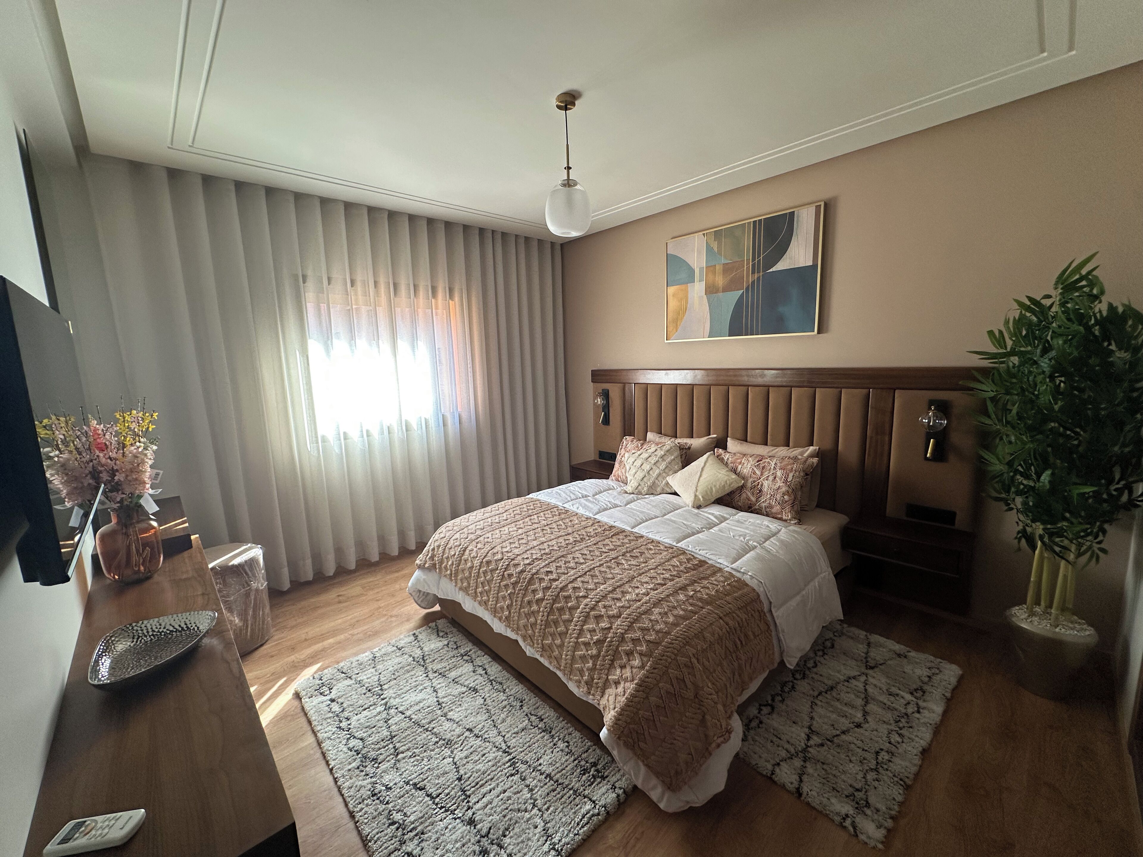 A modern hotel room with a large bed, a bedside table, a nightstand, a potted plant, and a painting on the wall.