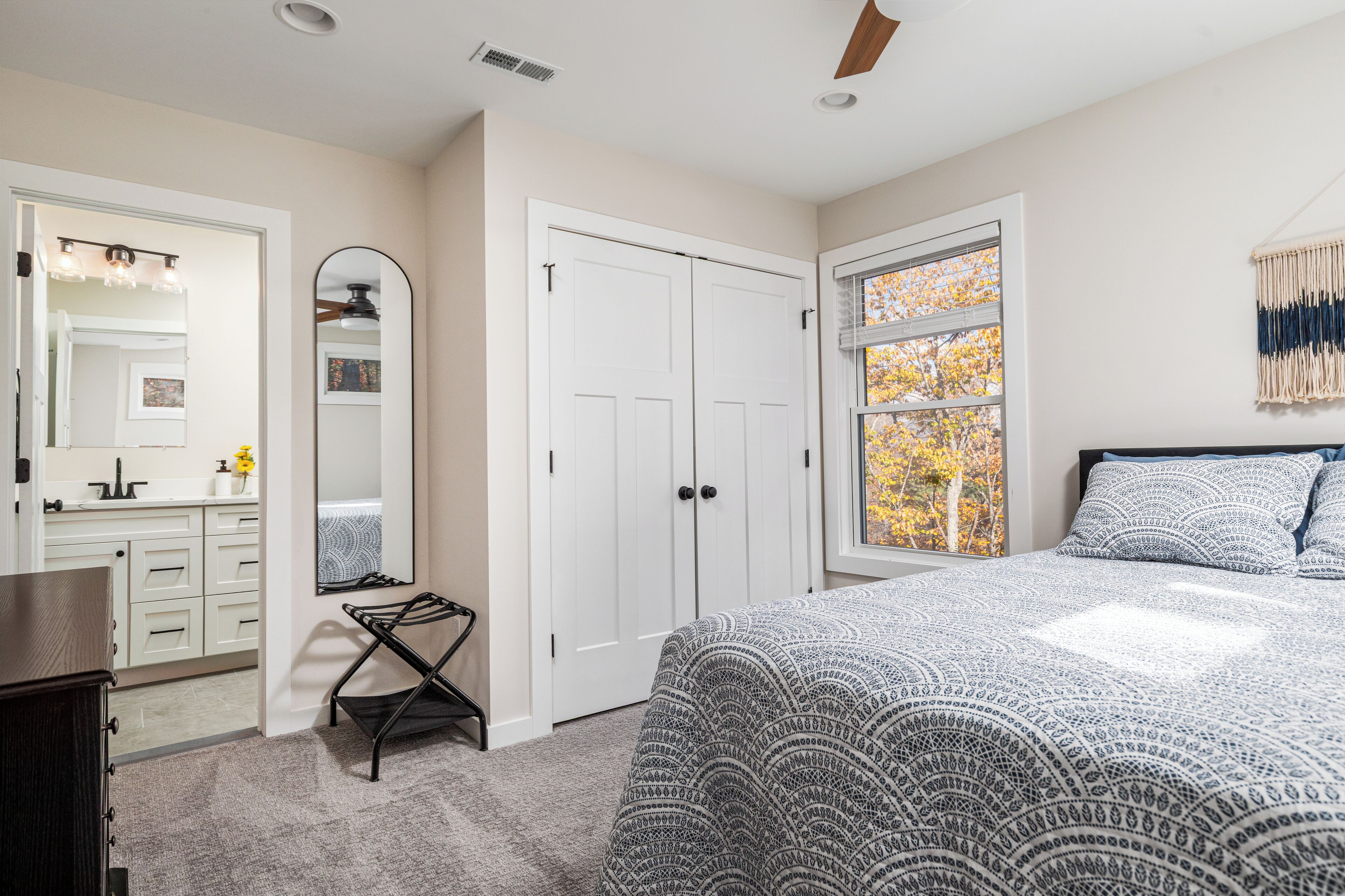 Une chambre à coucher avec un lit, une armoire, un miroir et une fenêtre donnant sur des feuilles d’automne.
