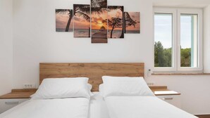 Un lit double avec du linge de lit blanc, une tête de lit en bois et une table de chevet avec une lampe. Il y a une fenêtre donnant sur un paysage verdoyant et un tableau représentant un coucher de soleil sur la mer.