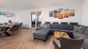 Un salon moderne avec un canapé d’angle gris, une table basse en bois sur laquelle sont disposés des fruits, et un coin repas avec une table et des chaises en bois.
