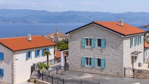 Un bâtiment en pierre avec des volets bleus et un toit rouge, entouré d’autres bâtiments, et offrant une vue sur la mer et les montagnes.