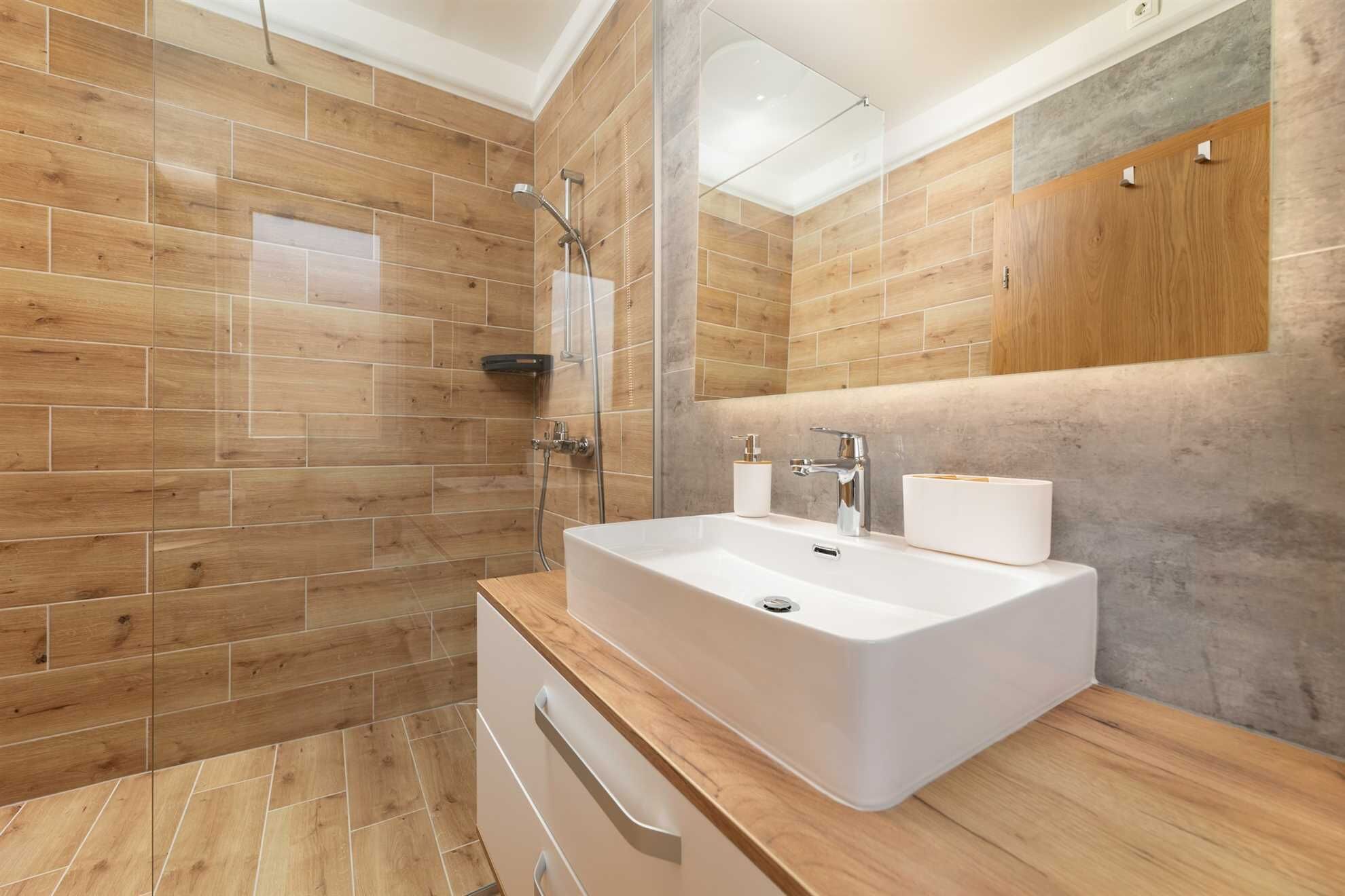 A modern bathroom with wooden flooring, a large white sink, a glass shower enclosure, and a wooden cabinet above the sink.