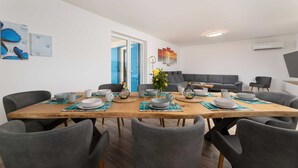 A dining area with a wooden table, grey chairs, and a grey sofa. There are wall art pieces and a window with blue shutters. - Villa Maja– Stylish Oasis with Heated Pool & Sea View (Vrbnik (Krk))