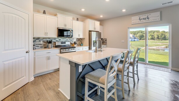 Una cocina moderna con armarios blancos, una isla central con taburetes de bar, un refrigerador de acero inoxidable y vistas a un paisaje verde a través de puertas correderas de vidrio grandes.