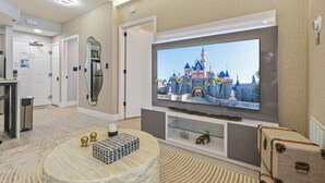 Habitación de hotel con una televisión de pantalla plana grande montada en la pared, una mesa de centro redonda con adornos, un aparador de vidrio debajo de la televisión y una maleta en el suelo.