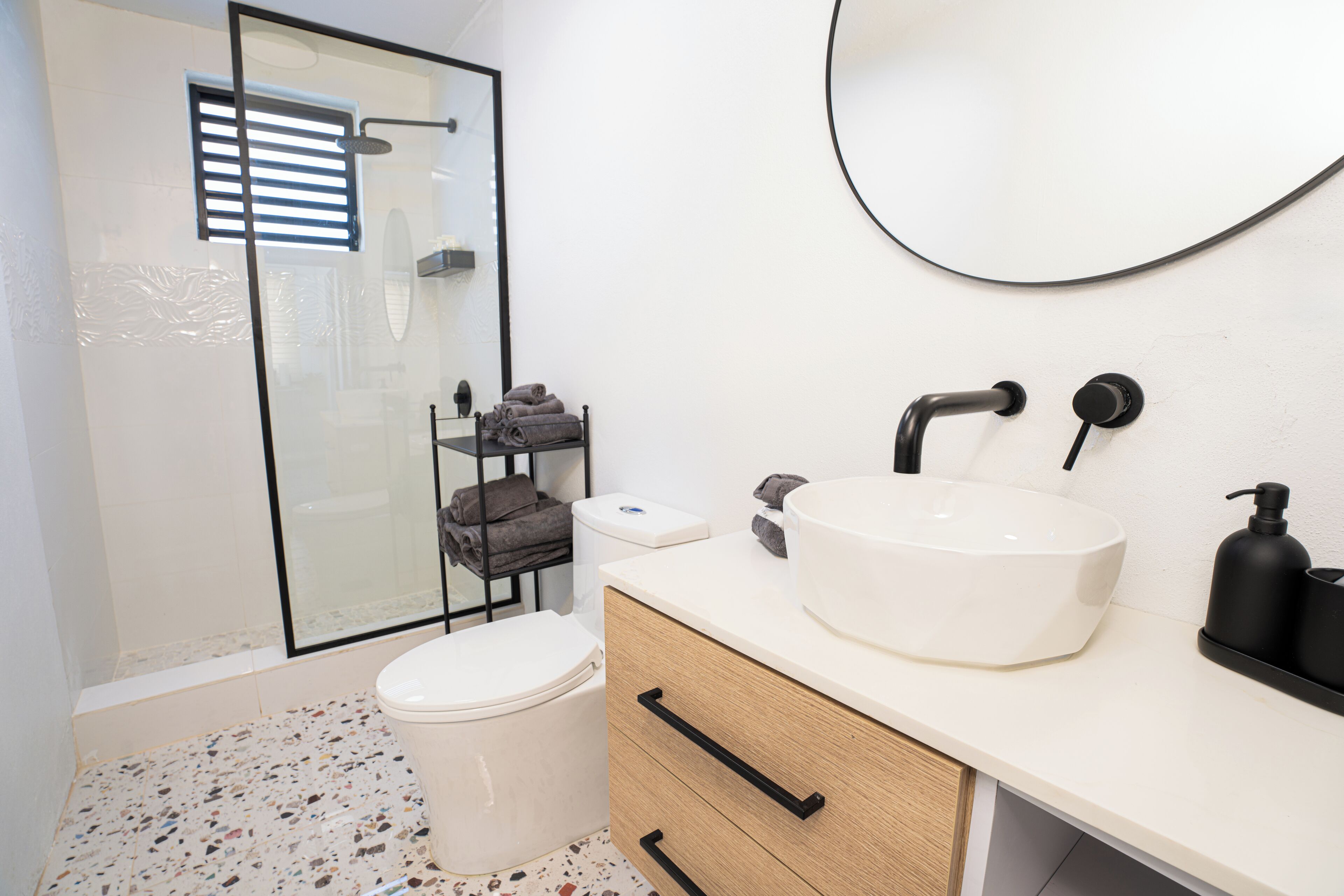 Une salle de bain moderne avec une douche à l’italienne en verre, un lavabo blanc avec un robinet noir et un meuble en bois doté de tiroirs.