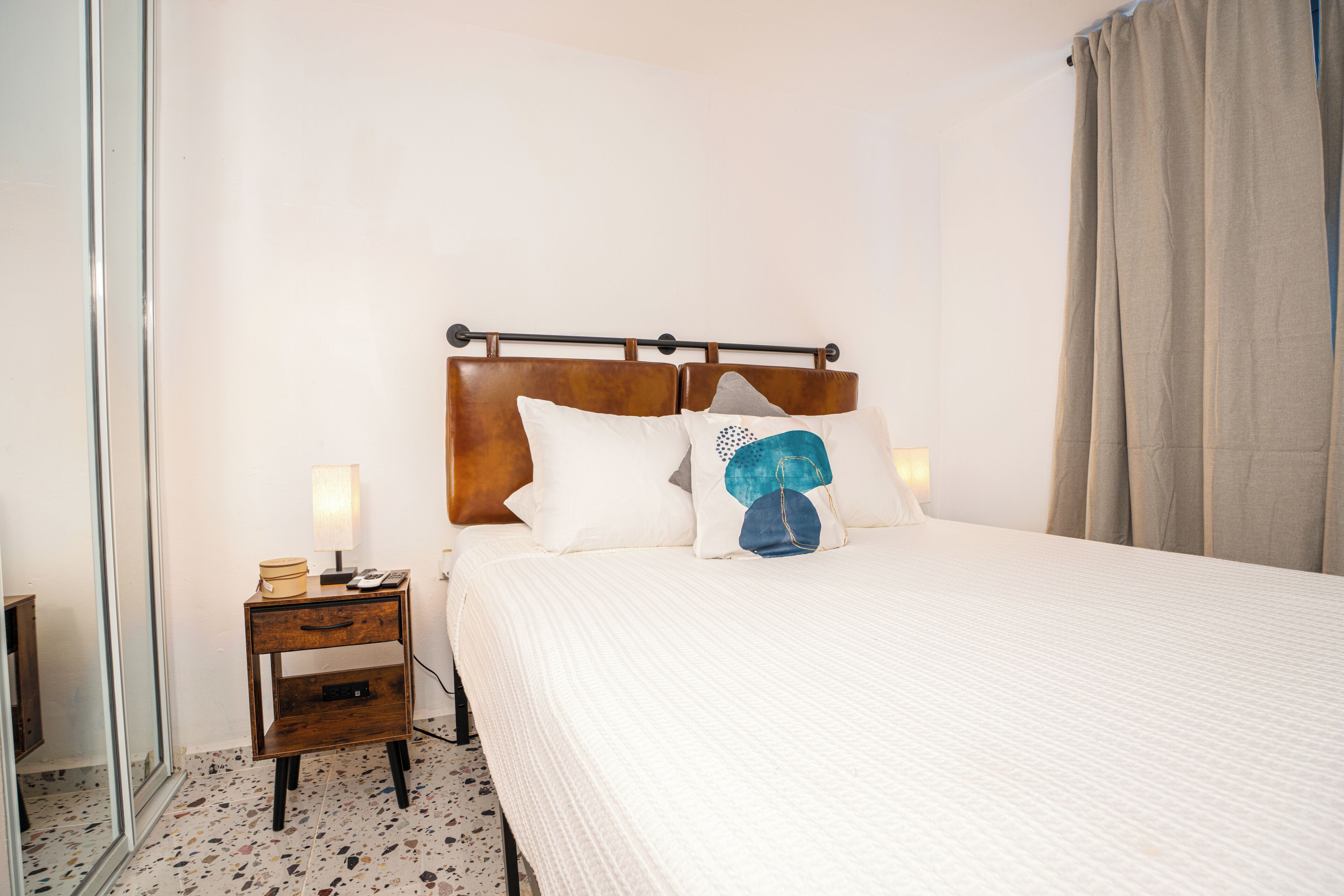 A neatly made bed with a wooden headboard, a bedside table with a lamp, and a mirror on the left.