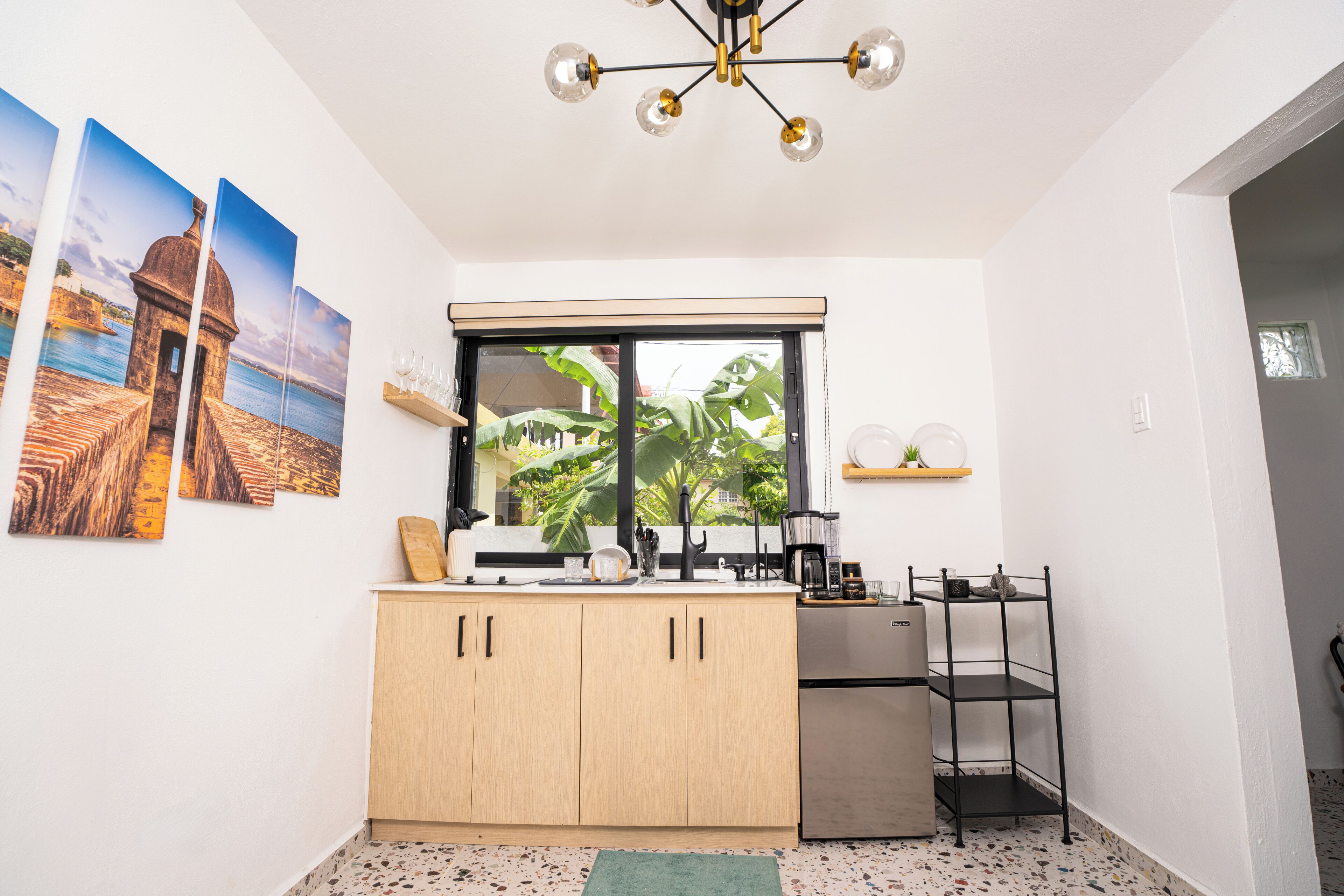 A compact kitchen with a window, wooden cabinets, a stainless steel refrigerator, and a black metal shelf.