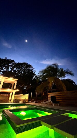A nighttime view of a swimming pool area with a moon in the sky, surrounded by palm trees and a wooden structure. - Garden of Eden Hotel (Vega Alta)