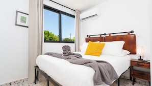 A hotel room with a large window, a bed with white linens, a wooden headboard, a bedside table with a lamp, and a rolled towel on the bed. - Garden of Eden Hotel (Vega Alta)