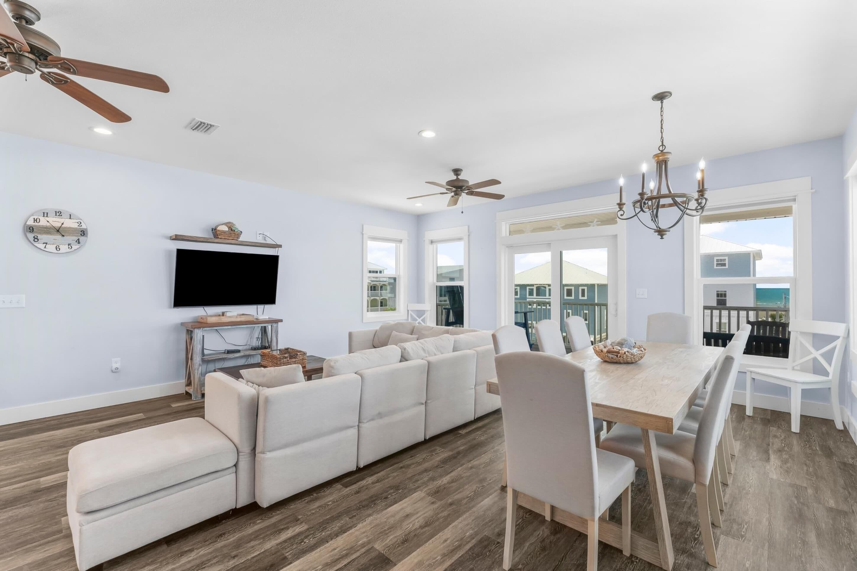 A modern living room with a sectional sofa, a dining table with chairs, a chandelier, ceiling fans, and a flat-screen TV mounted on the wall.