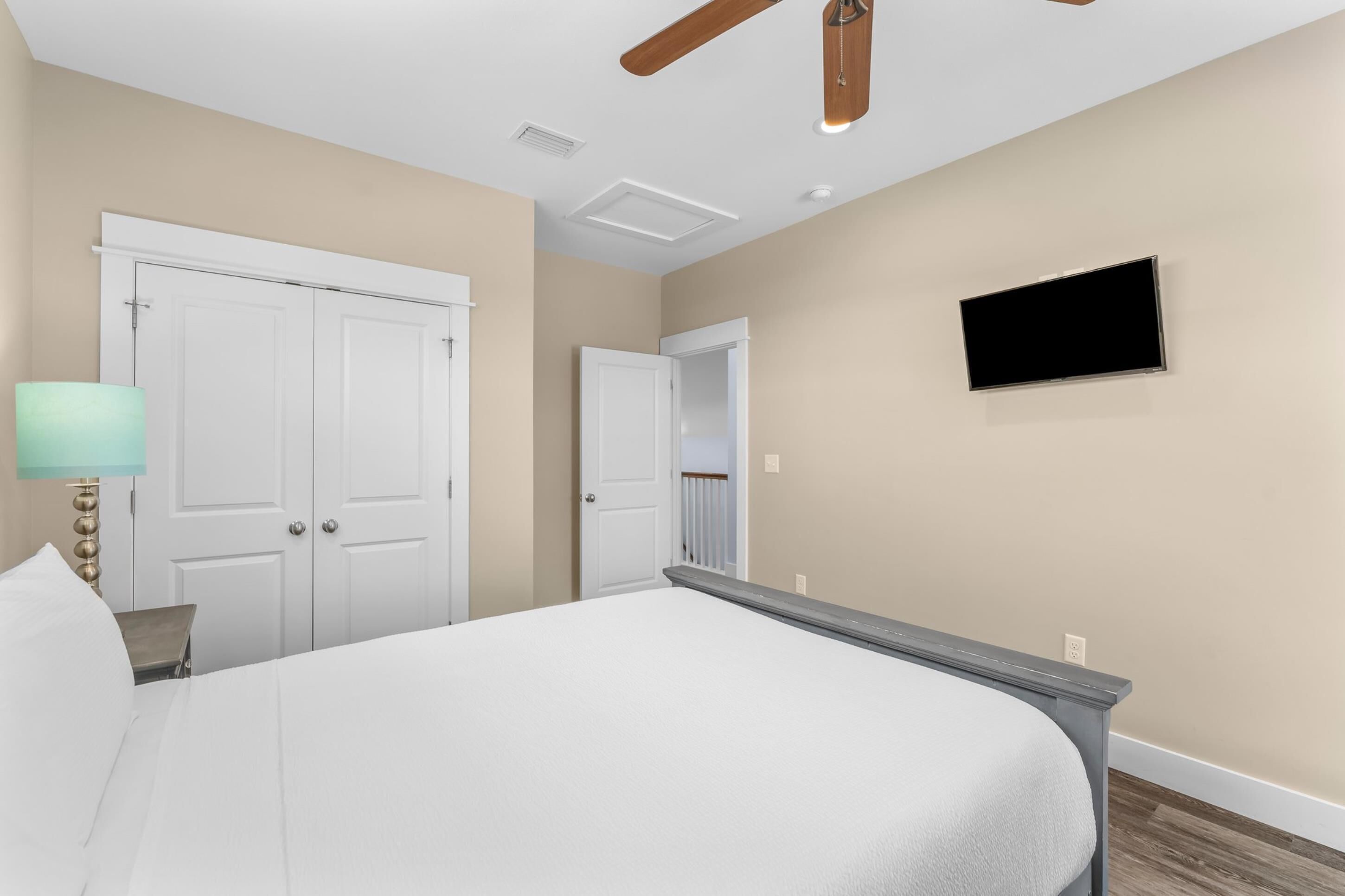 Un dormitorio con cama, ventilador de techo, televisor montado en la pared y una puerta que da a otra habitación.