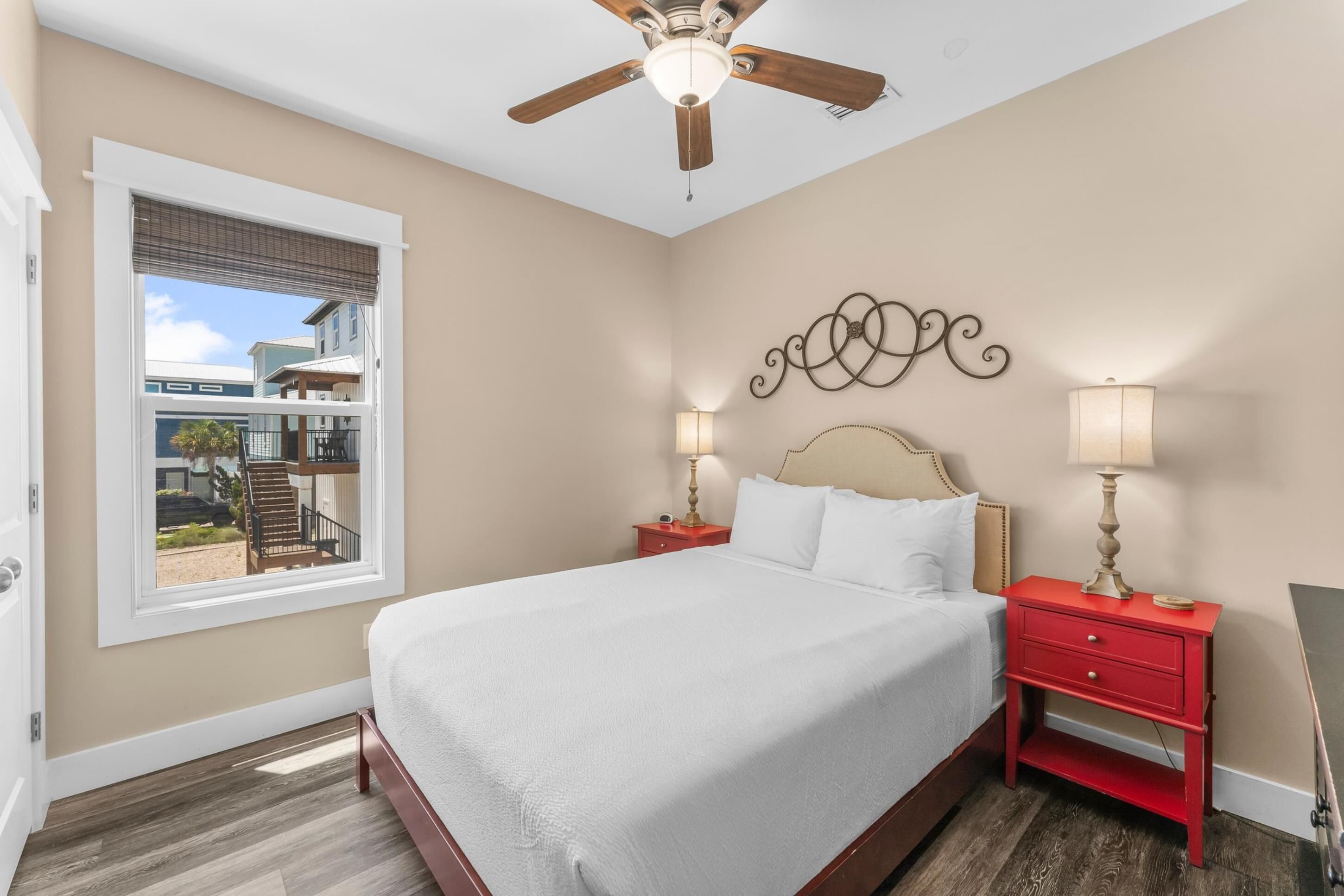A bedroom with a large bed, two bedside tables with lamps, a ceiling fan, and a window with a view of a building outside.