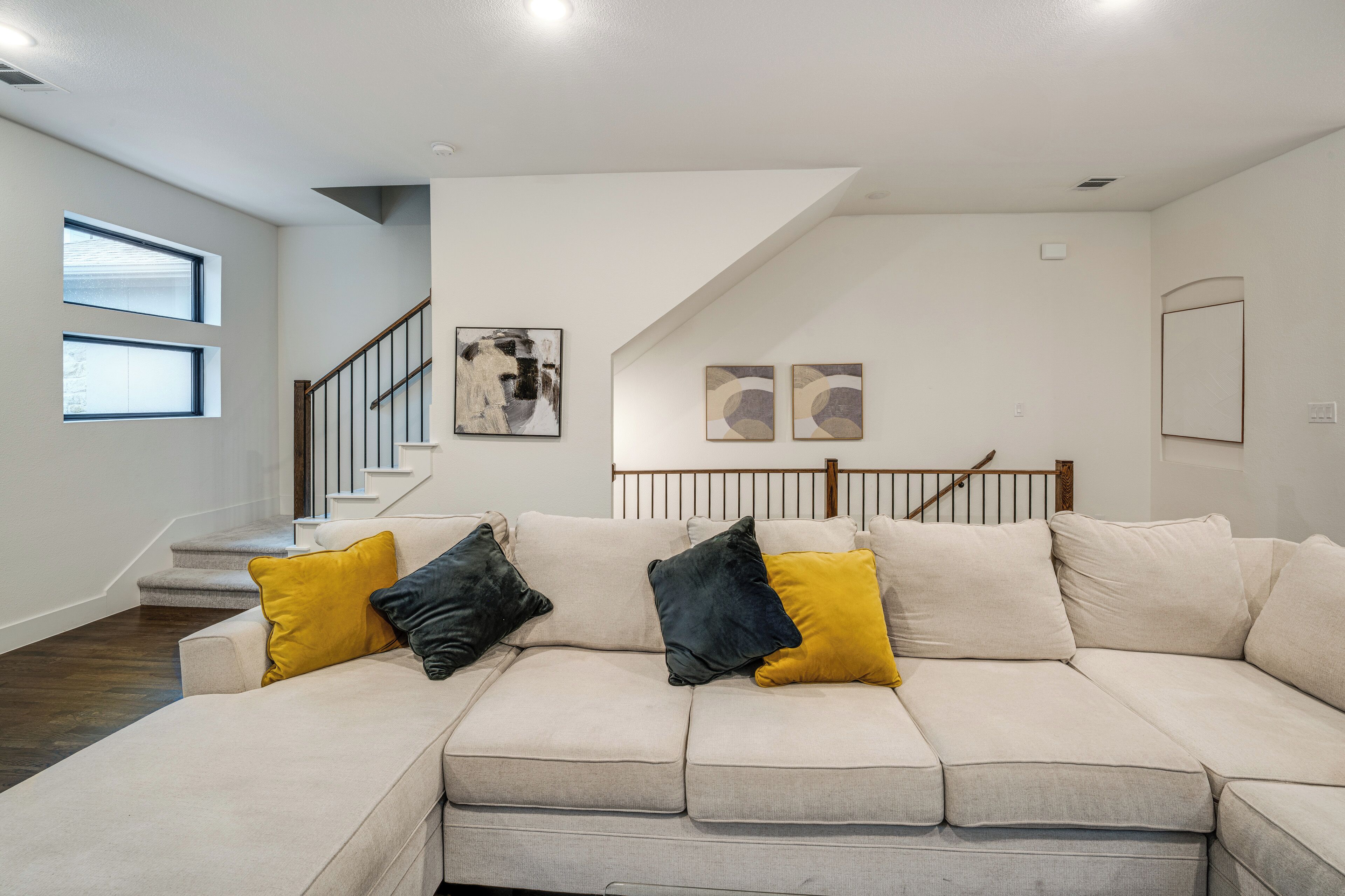 A modern living room with a beige sectional sofa, two windows, a staircase, and abstract wall art.