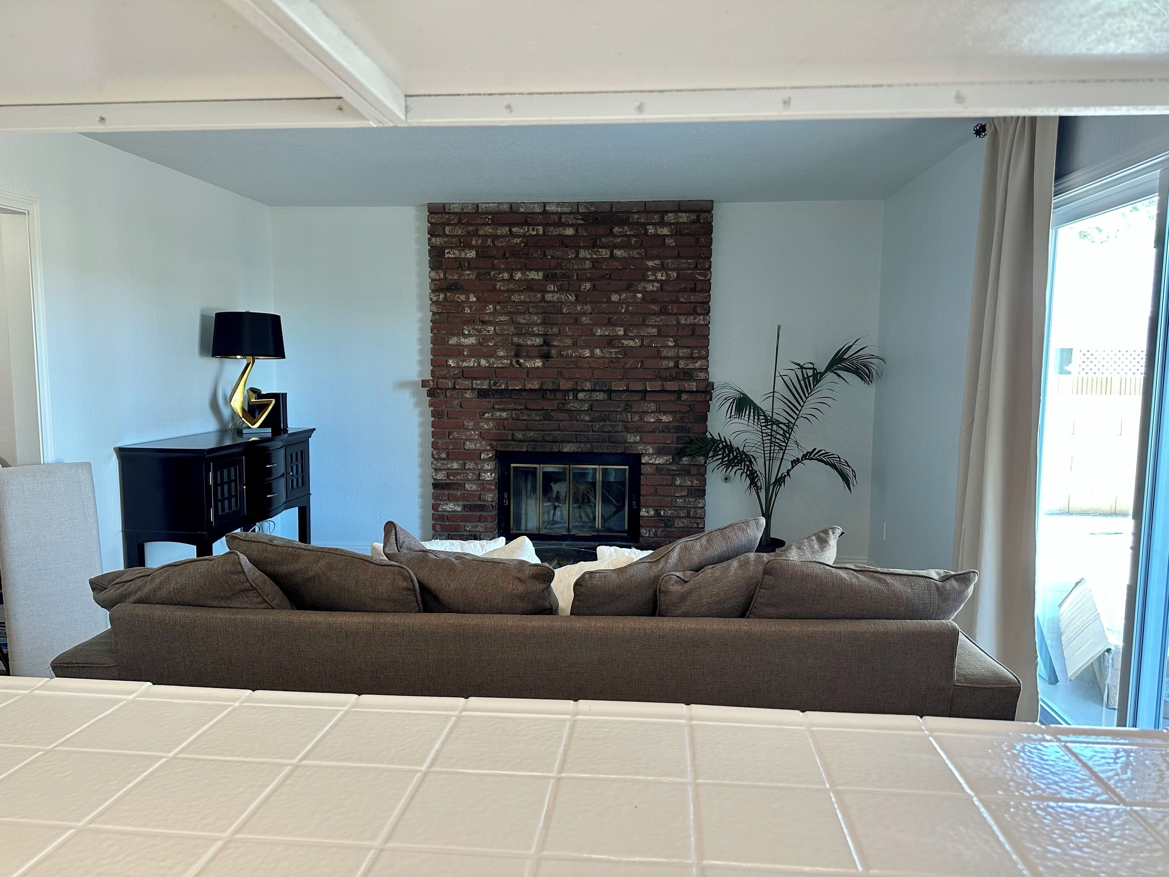 A living room with a brick fireplace, a sofa, a side table with a lamp, and a potted plant.