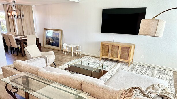 A modern living room with a sectional sofa, a glass coffee table, a wooden cabinet, a TV mounted on the wall, and a dining area with chairs and a table. - 4BR/2.5BA House in Delightful Lancaster with WiFi, AC and Large Yard (Lancaster)