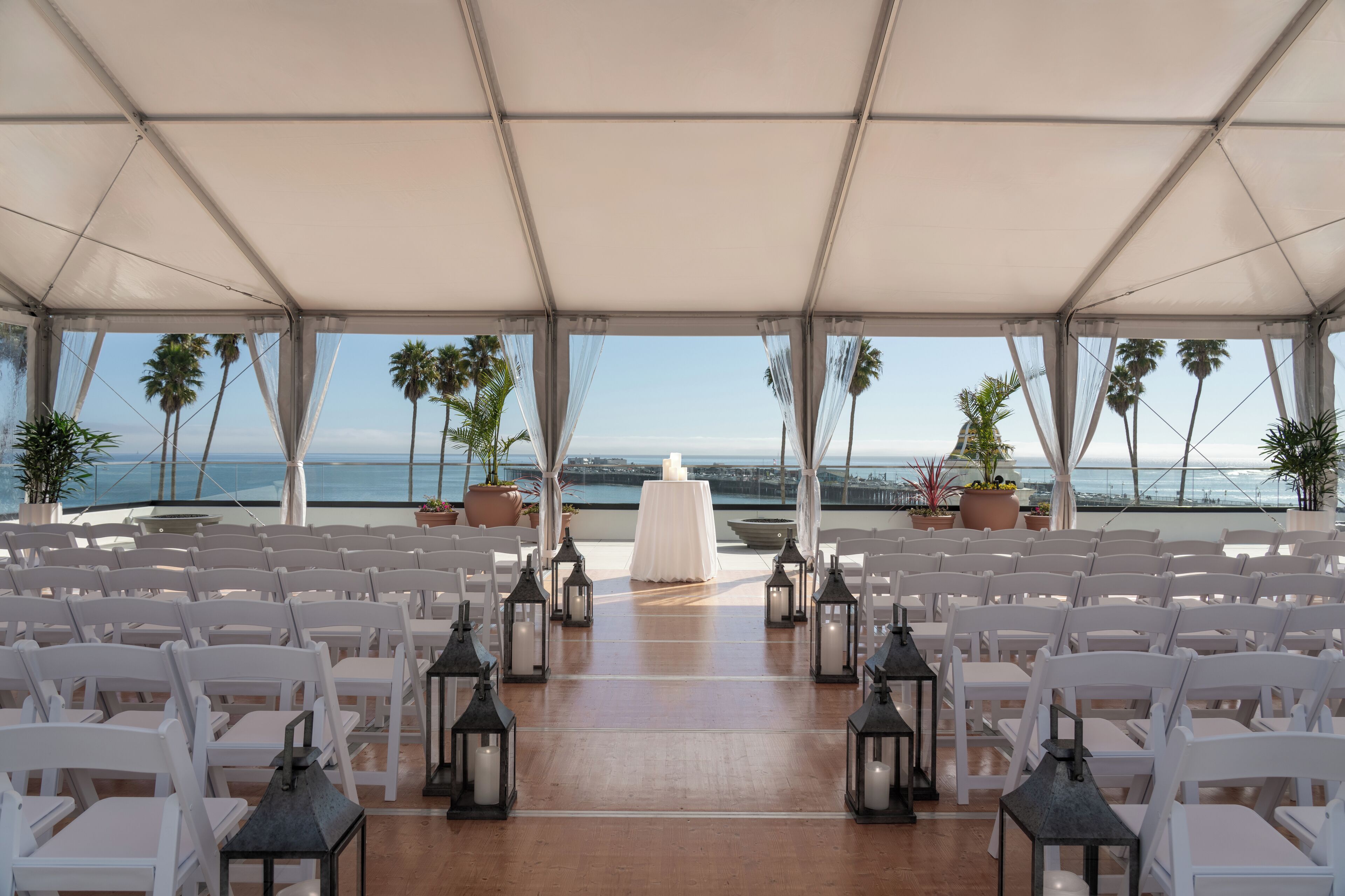 An outdoor wedding ceremony setup with white chairs and a white altar, surrounded by potted plants and overlooking a scenic ocean view.