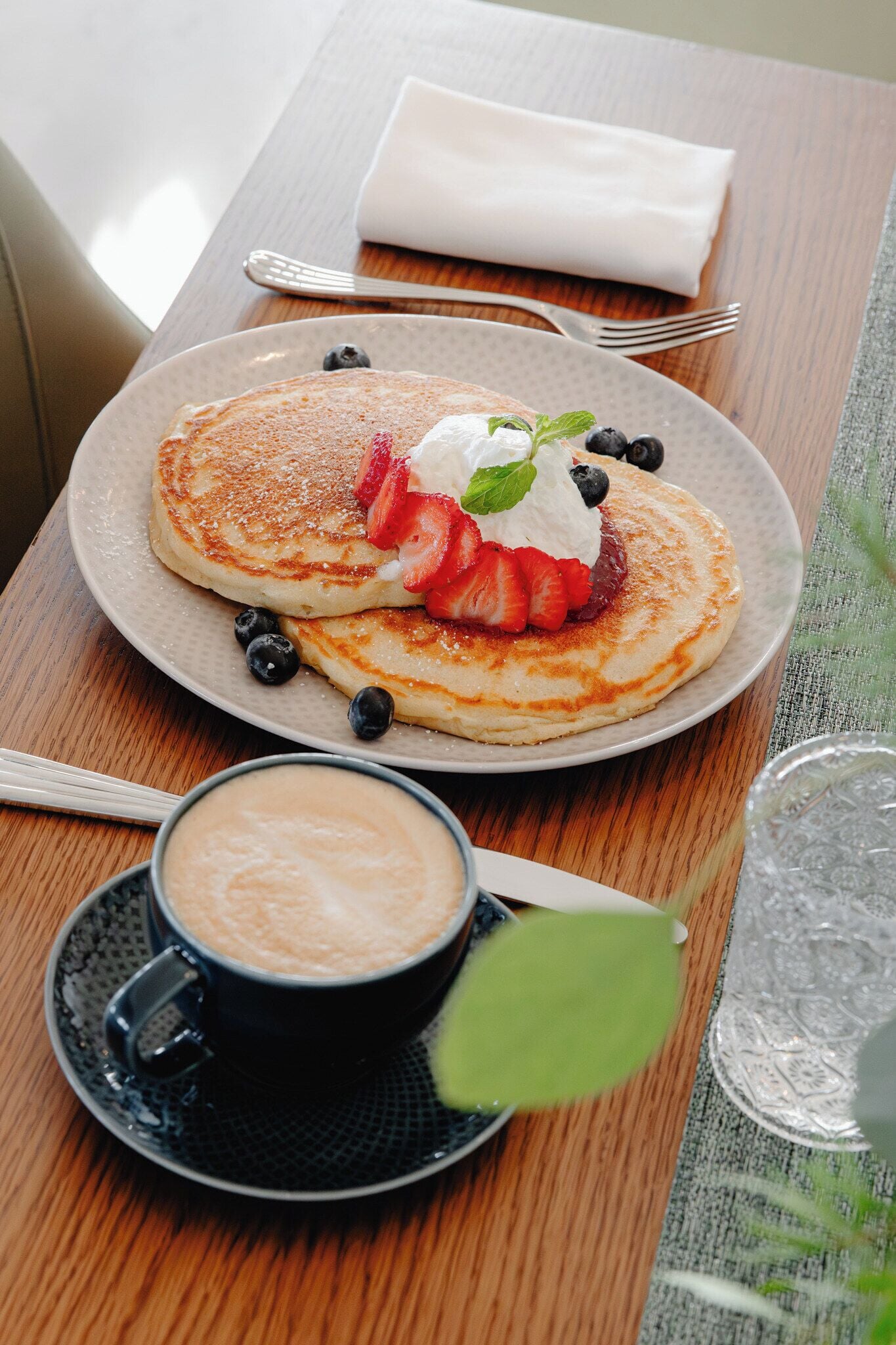 A breakfast setting with pancakes, strawberries, blueberries, whipped cream, and a cup of coffee on a wooden table.