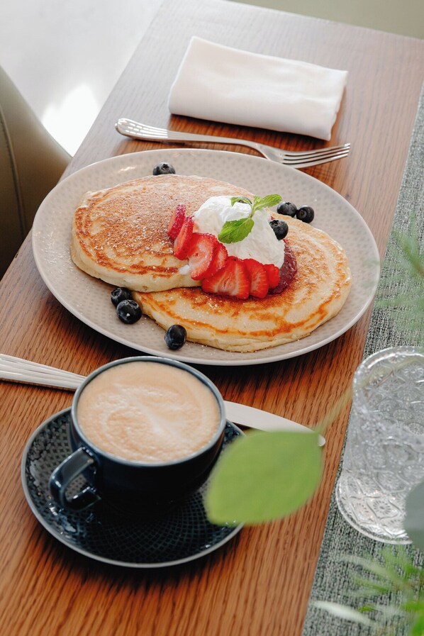 Desayuno con tortitas, fresas, arándanos, crema batida y una taza de café sobre una mesa de madera.