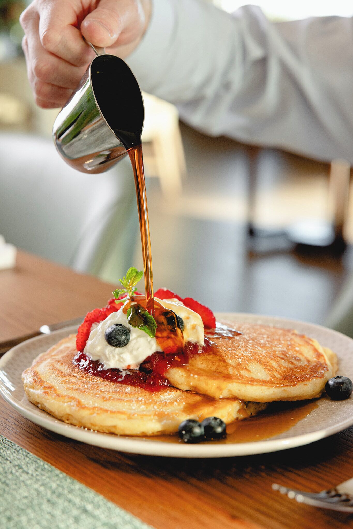 A hand pouring syrup over pancakes with berries and whipped cream on a plate.