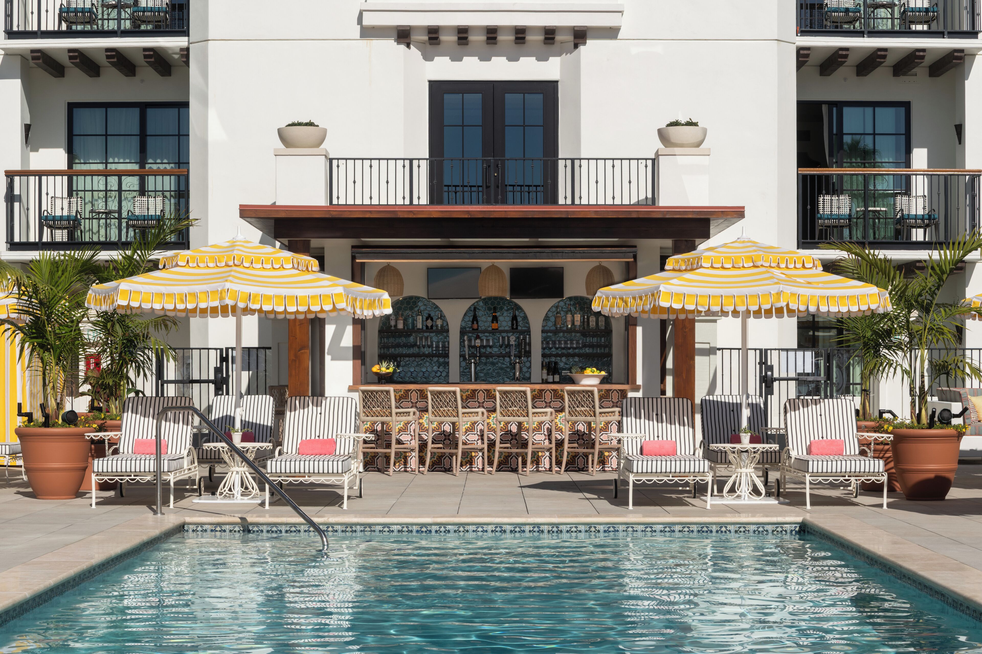 A pool area with lounge chairs and umbrellas, a bar with a tiled backsplash, and a building with balconies.
