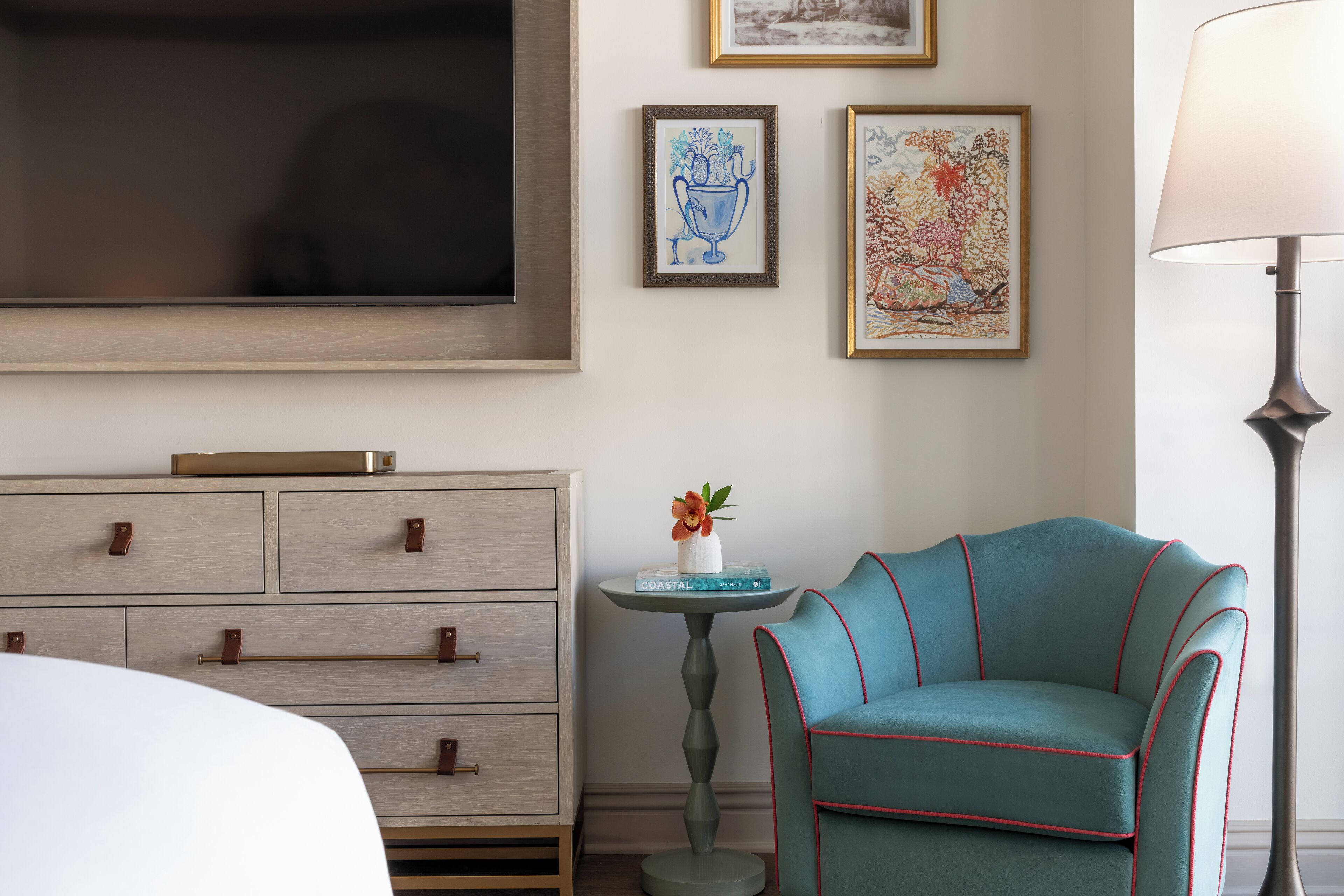 A hotel room with a bed, a dresser, a teal armchair, a small table with a vase, and framed artwork on the wall.