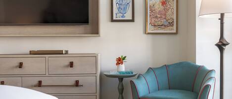 A hotel room with a bed, a dresser, a teal armchair, a small table with a vase, and framed artwork on the wall.