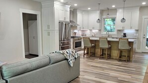 Una cocina moderna con armarios blancos, una isla central con taburetes de bar, un ventilador de techo y un área de comedor con sofá.