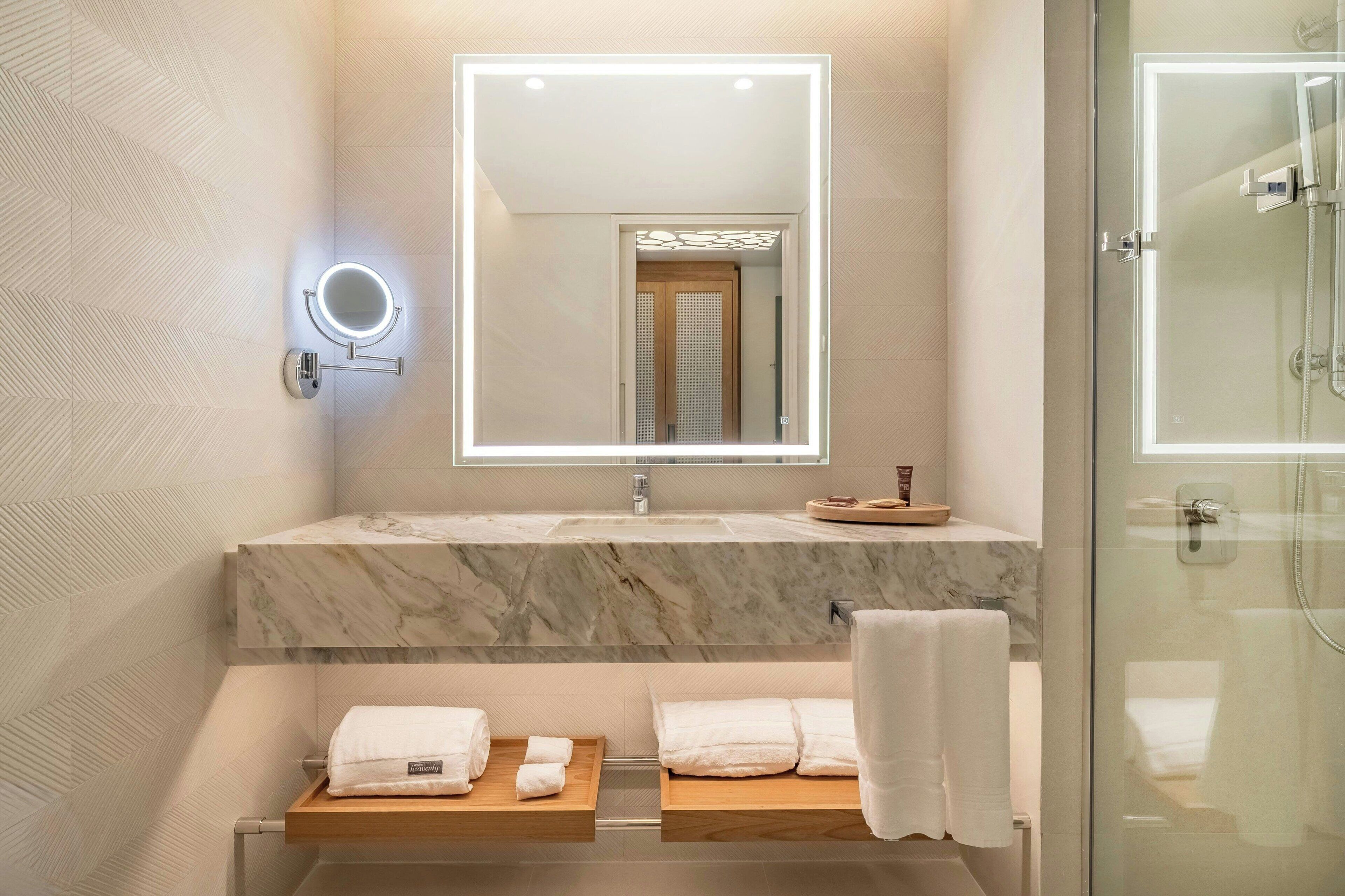 A modern bathroom with a marble sink, a mirror with integrated lighting, a glass shower enclosure, and neatly arranged towels.