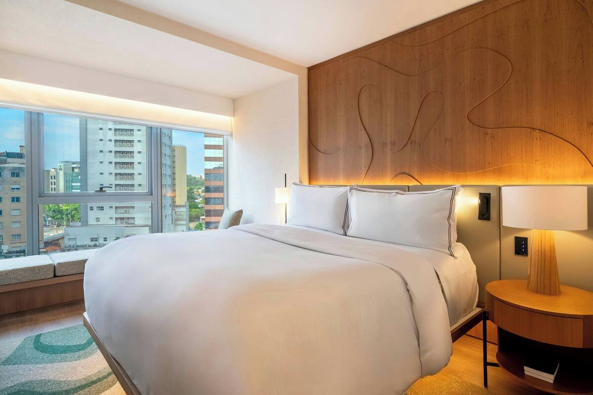 Ein modernes Hotelzimmer mit einem großen Bett, einer Wandvertäfelung aus Holz, Blick auf die Stadt und einem Nachttisch mit Lampe.