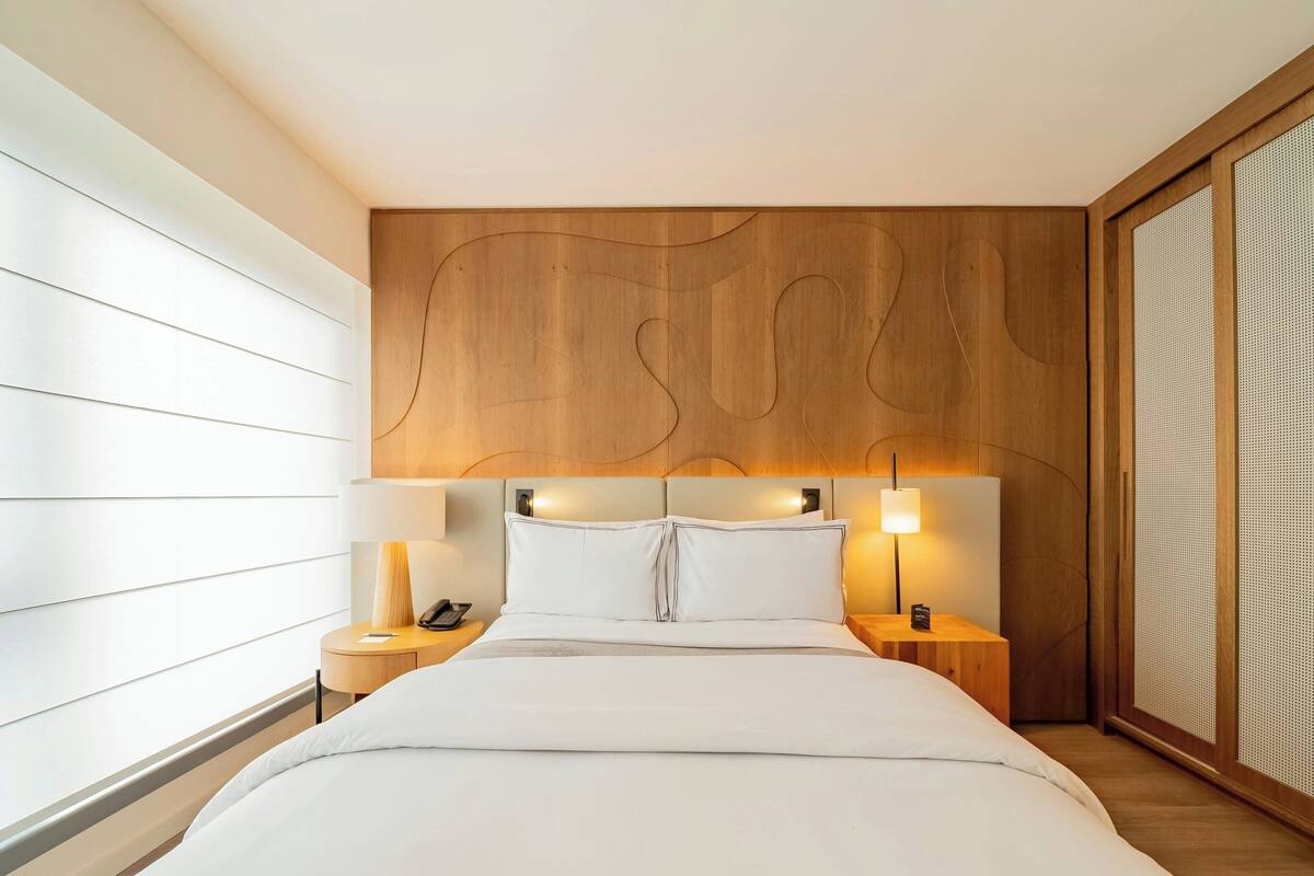 Ein modernes Hotelzimmer mit einem Holzkopfteil, einem großen Bett mit weißer Bettwäsche, zwei Nachttischen mit Lampen und einem großen Fenster mit Jalousien.