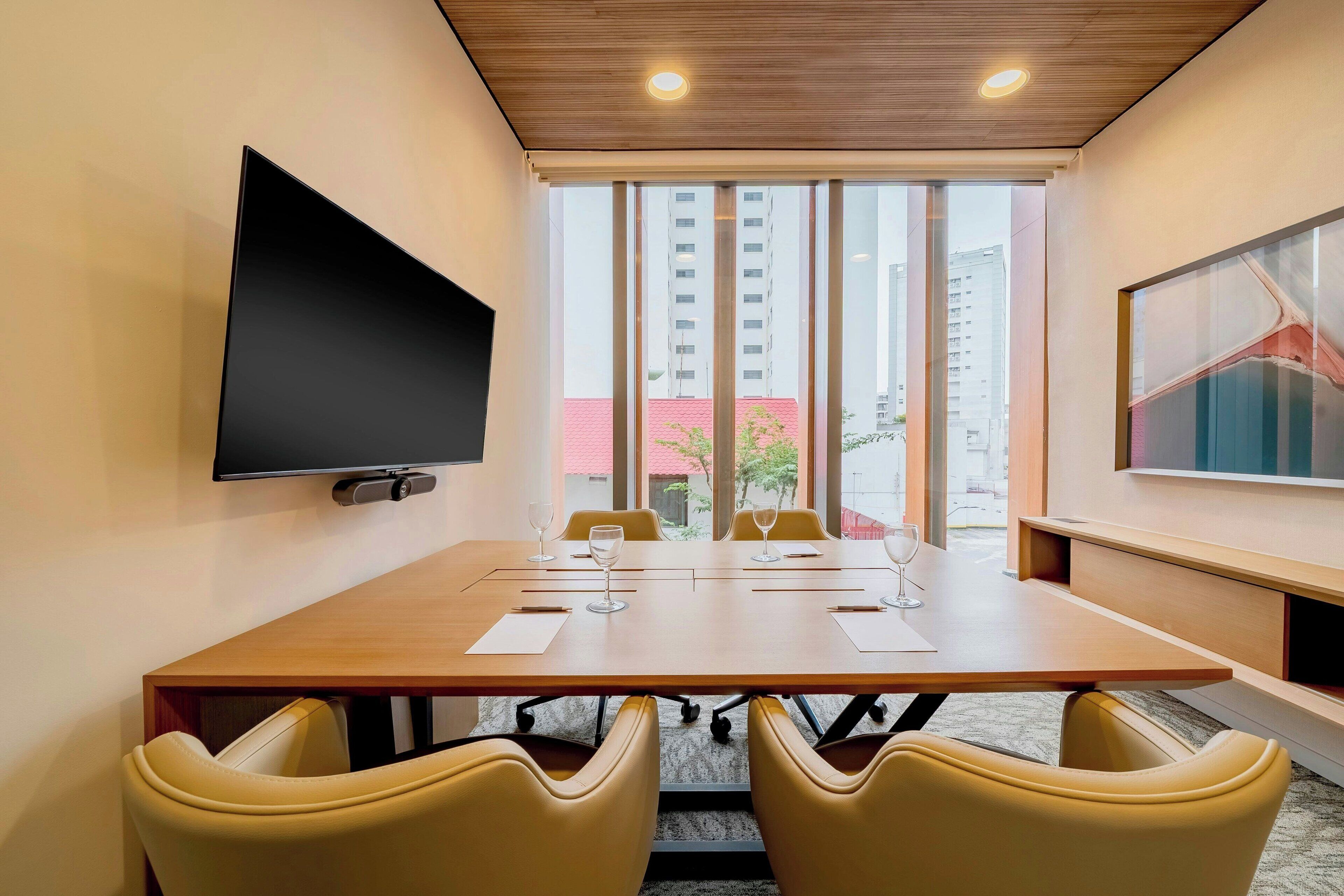 Un salón de conferencias con una larga mesa, sillas amarillas, un televisor de pantalla plana y ventanas amplias con vista a la ciudad.
