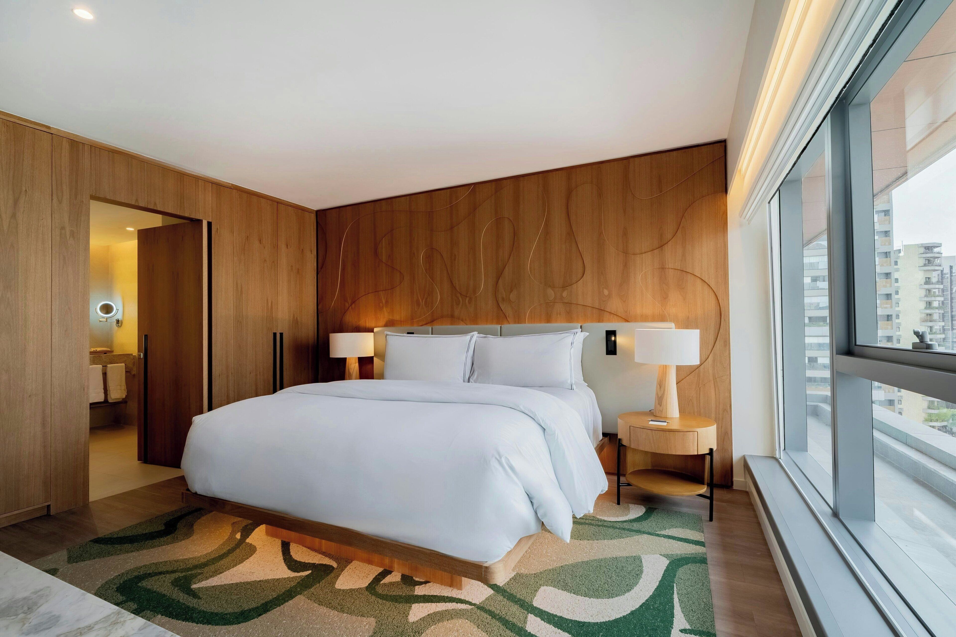 Une chambre d’hôtel moderne avec un grand lit, des panneaux de bois au mur, une vue à travers une grande fenêtre et une salle de bain visible par une porte ouverte.