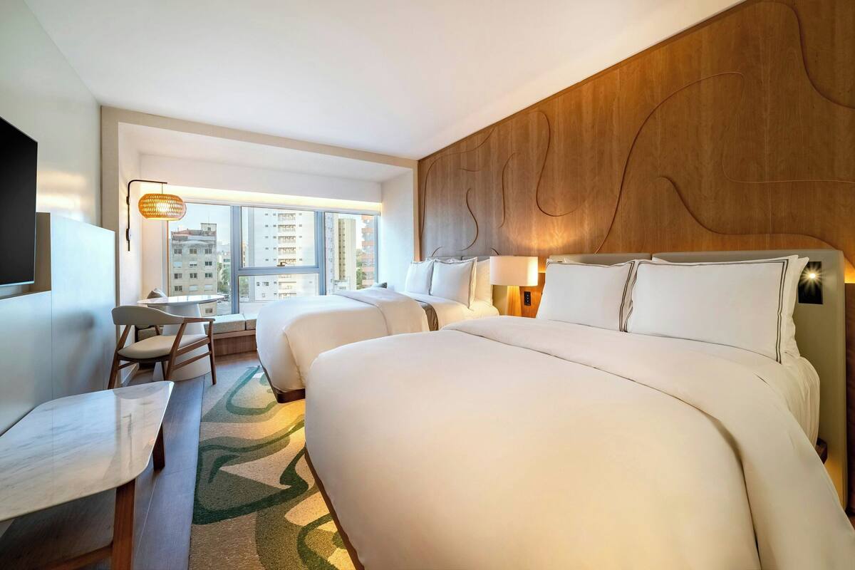 Ein Hotelzimmer mit einem großen Bett, zwei Nachttischen, einem Schreibtisch mit Stuhl und Blick auf die Stadt durch große Fenster.