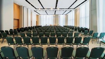 Un salón de conferencias con filas de sillas verdes, un escenario con un atril y ventanas grandes con persianas.