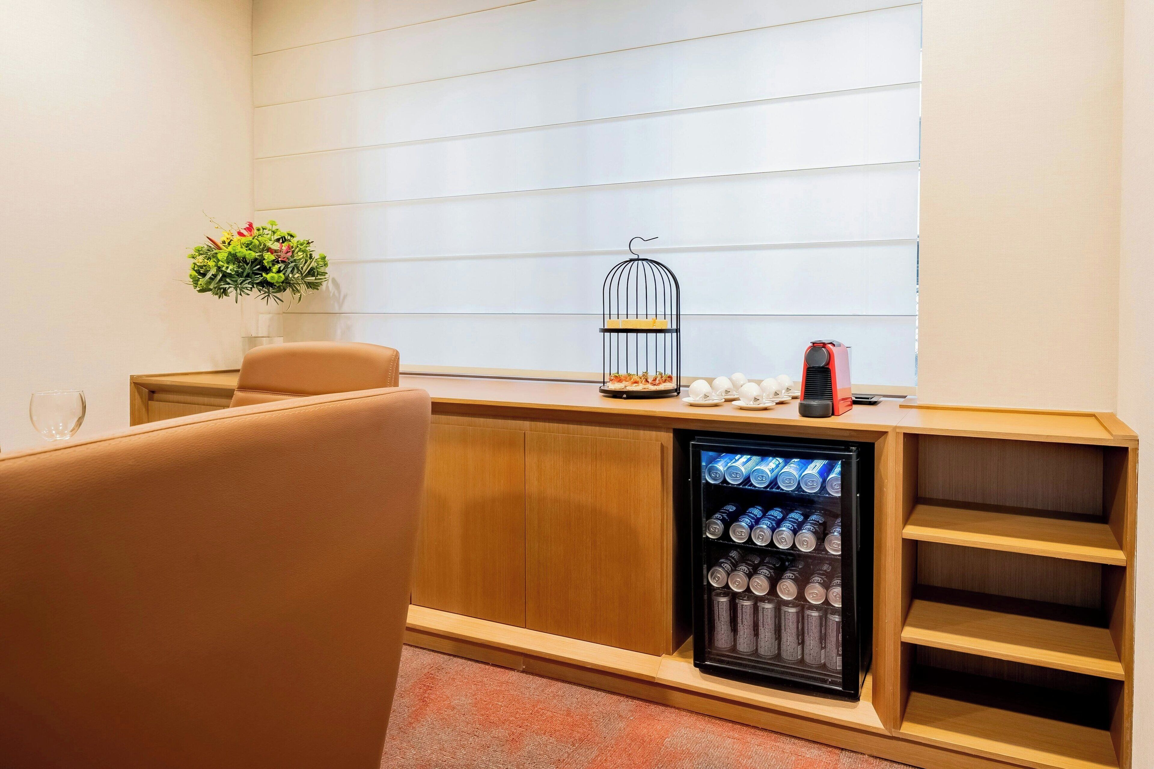 Habitación de hotel con minibar, un escritorio de madera, una silla, una mesita pequeña, una jaula decorativa y una planta en maceta.
