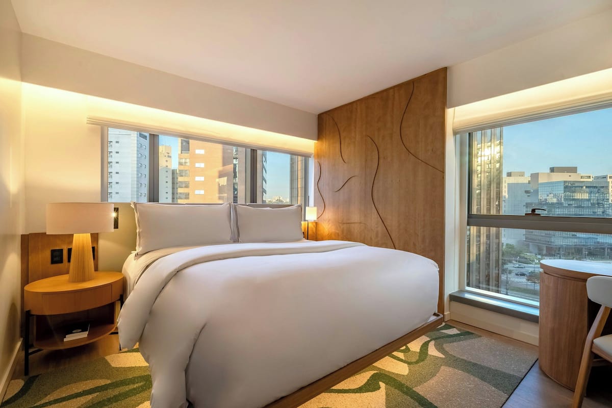 Ein Hotelzimmer mit einem großen Bett, einem Nachttisch mit Lampe, einem kleinen runden Tisch und Blick auf die Stadt durch große Fenster.