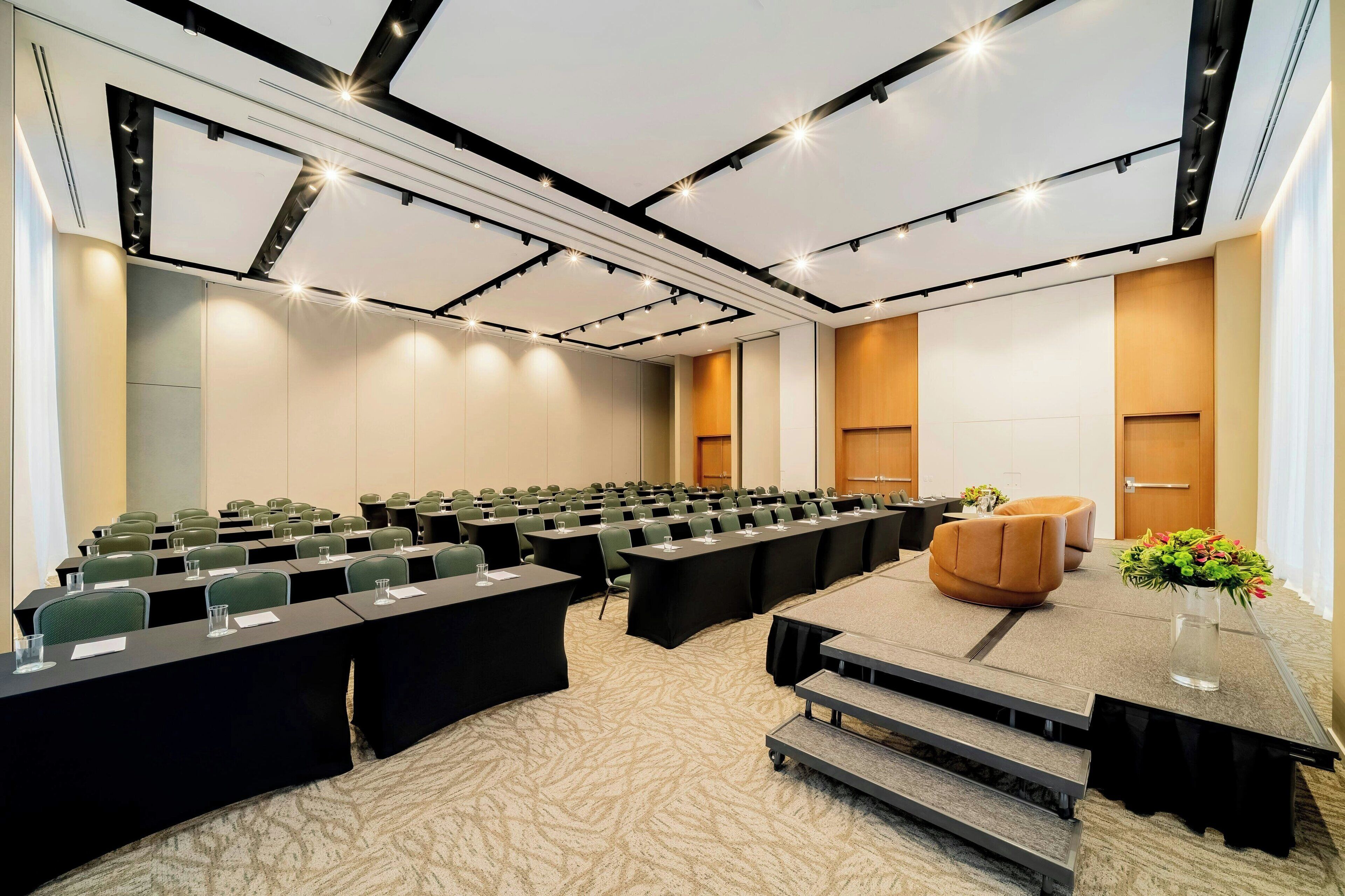 A conference room with rows of tables and chairs, a stage with a podium, and a carpeted floor.