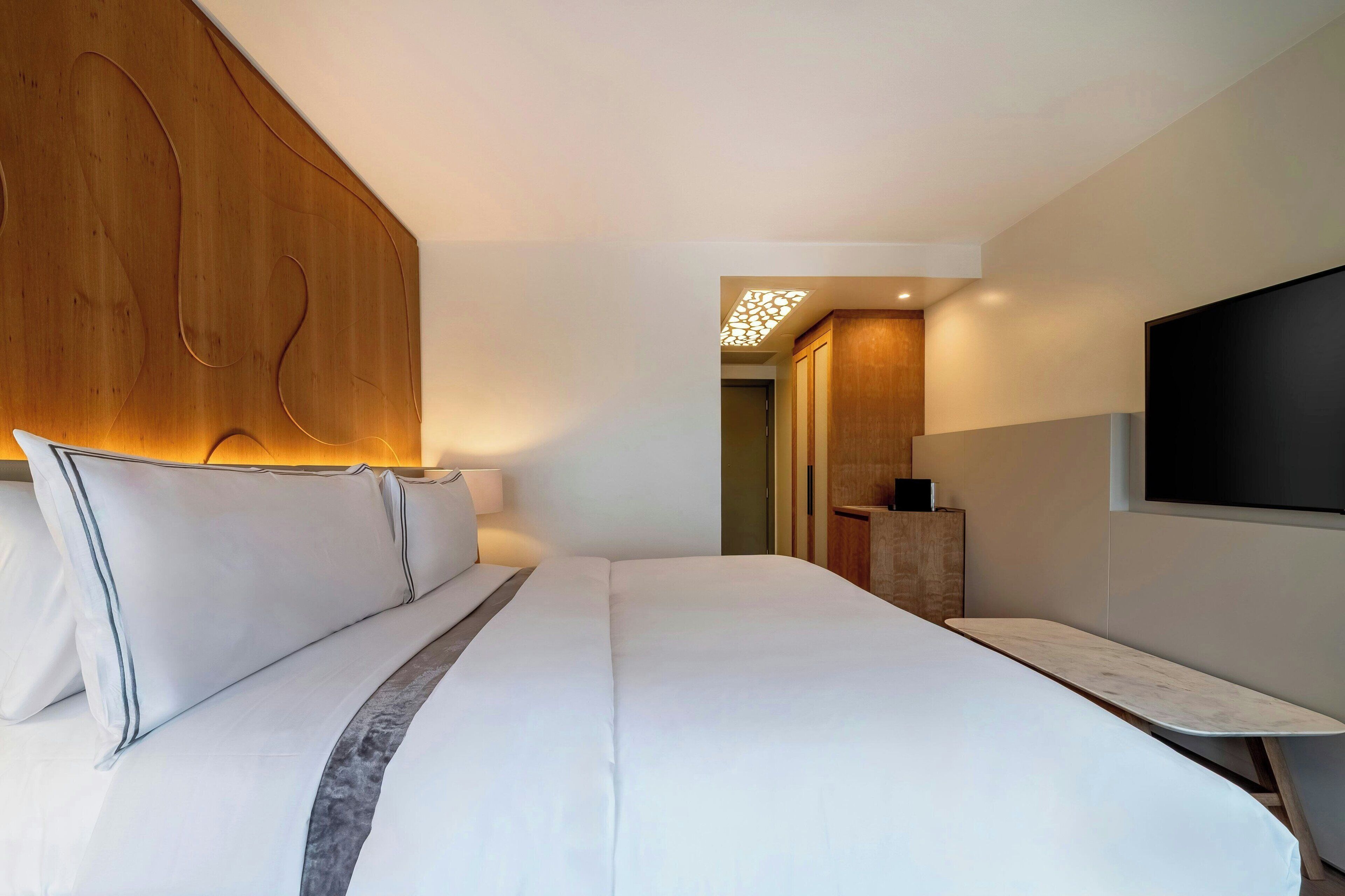 A modern hotel room with a large bed, wooden headboard, a flat-screen TV mounted on the wall, and a small desk area.