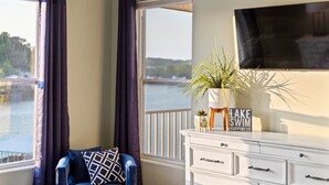 A room with a view of a body of water, a chair with a patterned cushion, a white dresser, and a sign that reads "LAKE SWIM HAPPINESS".