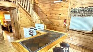 Interior de una cabaña de madera con una barra central tipo isla, pisos de madera, una escalera y un candelabro.