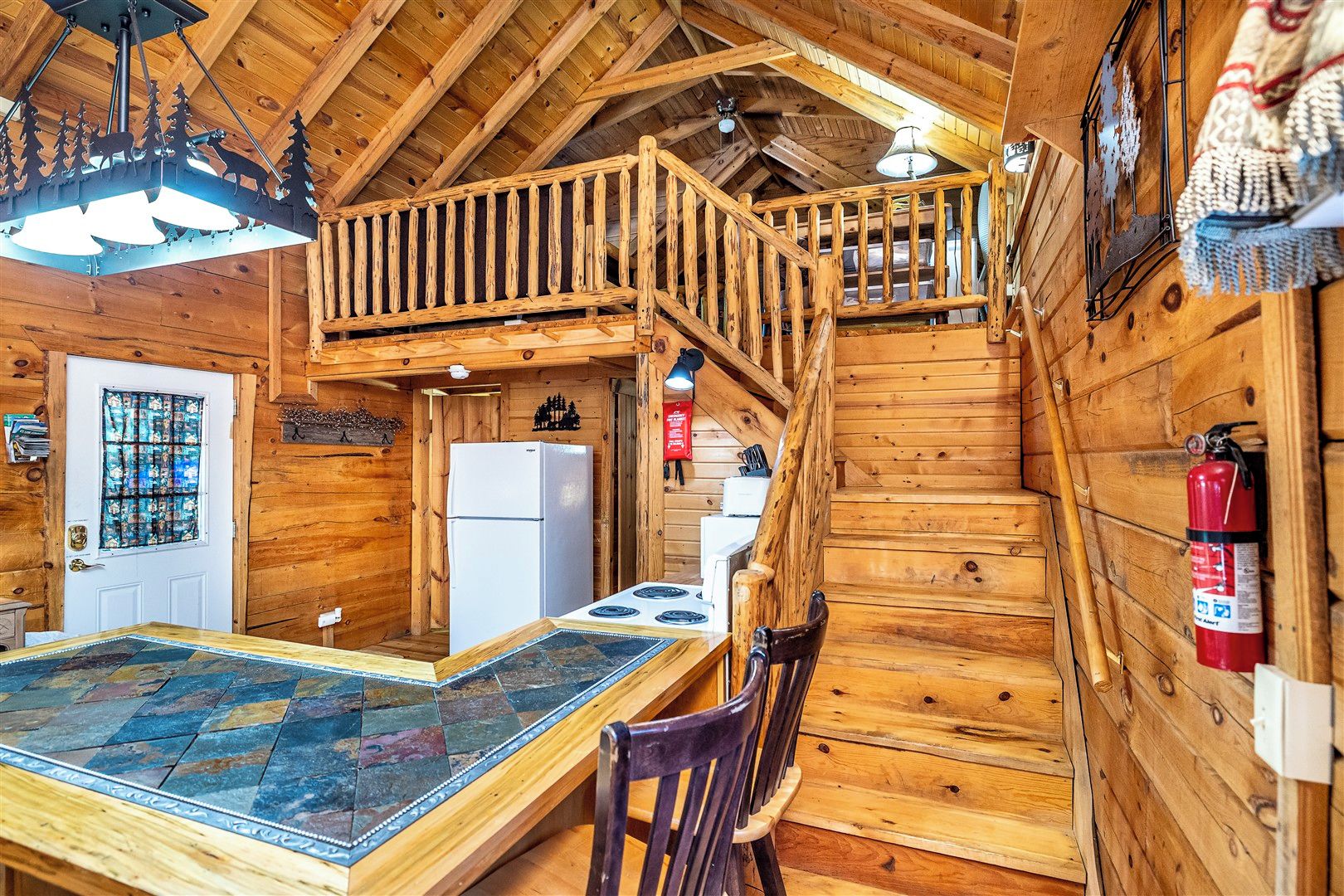 L’intérieur d’un chalet en bois comprend une table à manger, des chaises, un coin cuisine, un escalier et un luminaire au plafond.