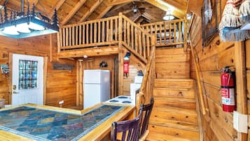 A wooden cabin interior with a dining table, chairs, a kitchen area, a staircase, and a ceiling light fixture.