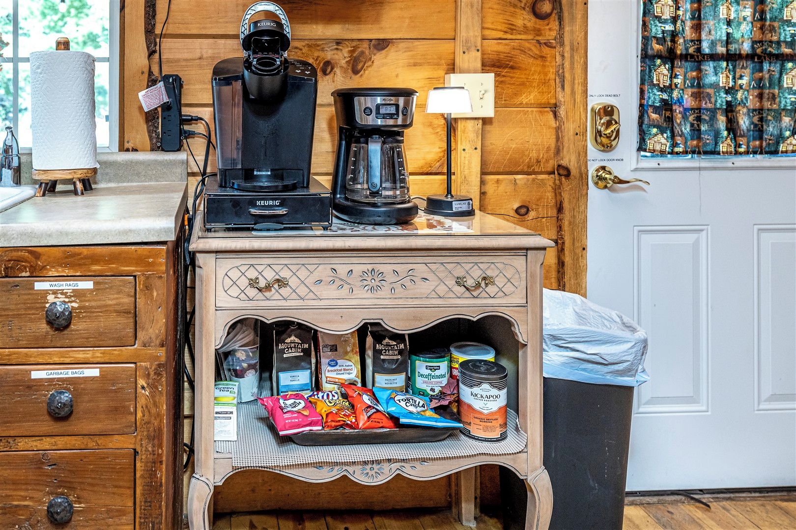 A kitchenette with a coffee maker, a kettle, and a coffee pot on a wooden countertop. Below, there are storage drawers labeled for wash rags and garbage bags.