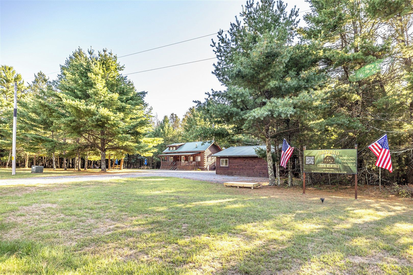 A cabin with a porch and a small adjacent building, surrounded by trees and a grassy area.