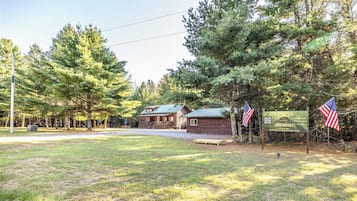 A cabin with a porch and a small adjacent building, surrounded by trees and a grassy area.