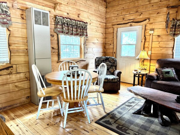 L’intérieur d’un chalet accueillant, avec une table à manger en bois, des chaises blanches, un fauteuil en cuir, un canapé et une lampe.