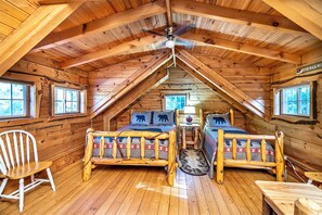 A cozy cabin with a sloped ceiling, wooden walls, two beds with matching headboards, a white chair, and a small table with a lamp. - Charming Log Cabin Fully Stocked 1.5 Garage Sleeps 7 UTV Route (Black River Falls)